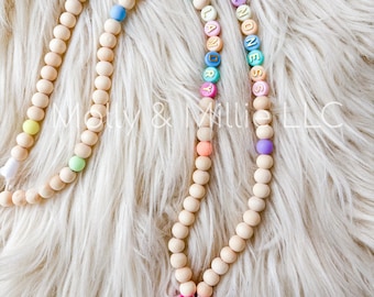 May include: A wooden beaded lanyard with a pink donut charm and a gold metal clip. The beads are in a rainbow pattern and spell out "SPRINKLE KINDNESS" and "SLANDRY".