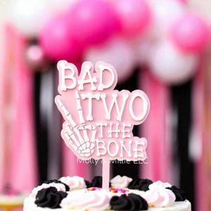 Pode incluir: Um topo de bolo rosa e branco com as palavras "BAD TWO THE BONE" e uma mão de esqueleto. O topo está colocado em um bolo decorado com glacê rosa e preto. O fundo apresenta balões e decorações rosa.