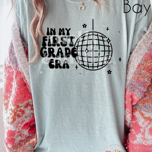 K&ouml;nnte beinhalten: Ein hellblaues T-Shirt mit dem Text "IN MY FIRST GRADE ERA" in schwarzen Buchstaben mit einer Discokugel-Grafik. Der Text ist in einem Retro-Schriftart-Stil. Das T-Shirt hat einen Used-Look.