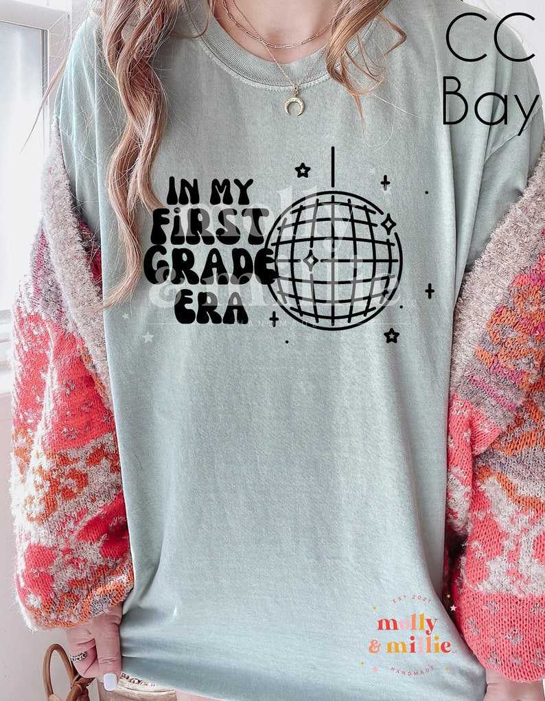 K&ouml;nnte beinhalten: Ein hellblaues T-Shirt mit dem Text "IN MY FIRST GRADE ERA" in schwarzen Buchstaben mit einer Discokugel-Grafik. Der Text ist in einem Retro-Schriftart-Stil. Das T-Shirt hat einen Used-Look.