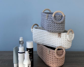 May include: Three rectangular crochet baskets in grey, white, and pink, stacked. Each basket has wooden handles. Skincare products are arranged to the left, including a tube, spray bottle, and bottles with black and white labels.