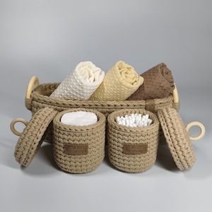 May include: A woven beige basket with rolled towels in cream, yellow, and brown. Two matching lidded containers hold cotton balls and cotton swabs. The containers have a label that reads "Cotton Family".