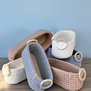 Op de afbeelding: Een verzameling handgemaakte gehaakte manden in verschillende maten en kleuren, waaronder wit, grijs en roze. Elke mand heeft houten handvatten en een stevig, geweven ontwerp. De manden zijn op een houten oppervlak geplaatst.