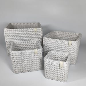 Puede incluir: Cuatro cestas de almacenamiento blancas tejidas de diferentes tamaños. Las cestas tienen forma rectangular con un diseño texturizado en bucle. Cada cesta tiene una pequeña etiqueta de color claro adjunta. Las cestas están dispuestas sobre una superficie blanca.
