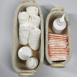 Peut inclure: Deux paniers tressés beiges avec des poignées en bois. L'un contient des serviettes blanches roulées et deux récipients. L'autre contient une bouteille avec une pompe et une pile de serviettes hygiéniques à motifs floraux orange.