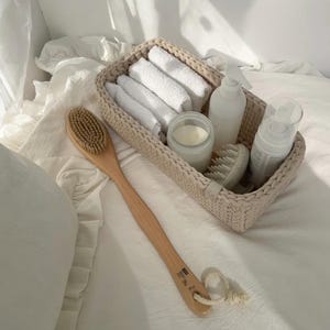 Op de afbeelding: Een rechthoekige beige gehaakte mand met vier gevouwen witte washandjes, een doorzichtige glazen pot met witte crème, een witte pompflacon, een ronde witte hoofdhuidmassager en een houten badborstel met een lange steel en een touwtje.