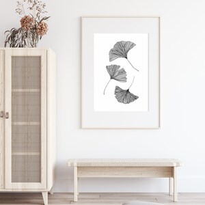 Puede incluir: Un dibujo en blanco y negro de cuatro hojas de ginkgo en un marco blanco colgado en una pared blanca. El marco est&aacute; sobre un banco de madera clara.