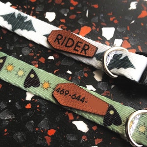 Puede incluir: Dos collares para perros con etiquetas de nombre de madera. El collar superior es blanco con murciélagos negros y la etiqueta dice "RIDER". El collar inferior es verde con murciélagos negros y estrellas y la etiqueta dice "469-644".