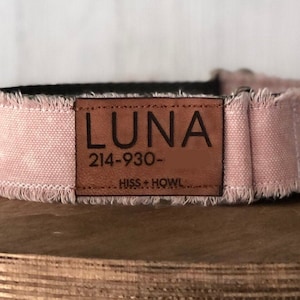 May include: A pink dog collar with a frayed edge and a black interior. A brown leather patch displays the name "LUNA" and a phone number. The collar is resting on a wooden surface.