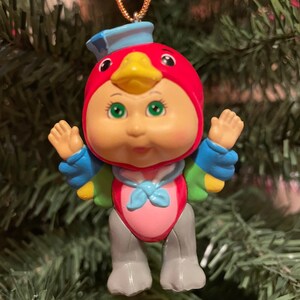 May include: A colourful Christmas ornament in the shape of a bird-faced doll. The ornament has a red head with a yellow beak, green eyes, and a blue hat. It has blue and green wings, a pink torso, and grey legs. A gold string is attached for hanging.