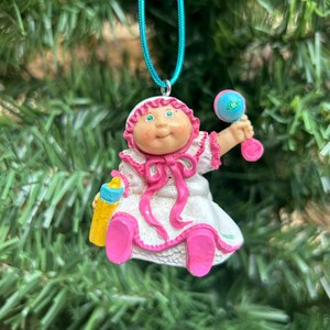 以下が含まれることがあります： キャベツ人形のクリスマスオーナメント。白いドレス、ピンクのアクセント、お揃いのボンネットを着た赤ちゃん人形が特徴です。人形は青いガラガラと黄色の哺乳瓶を持っています。ターコイズ色のコードから吊り下げられています。