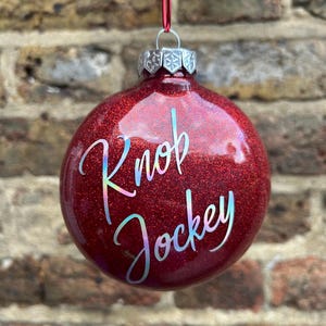 May include: Red glitter Christmas bauble with the words "Knob Jockey" written in silver glitter.