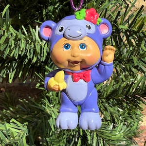 May include: A Cabbage Patch Kids ornament featuring a doll dressed in a purple monkey costume with a hood, pink inner ears, and a red bow tie. The doll has blue eyes, holds a yellow banana, and has a red flower on the hood. A purple cord is attached.