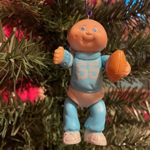 May include: A Cabbage Patch Kids Christmas ornament. The doll is wearing a light blue football uniform with the number 55 and holding a brown football. The ornament is suspended from a purple string.