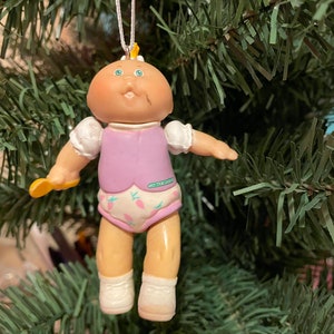 May include: A Cabbage Patch Kids doll Christmas tree ornament. The doll has a round head, blue eyes, and a small mouth. It wears a lavender vest, a diaper with carrot designs, and white shoes. The doll is holding a yellow object.