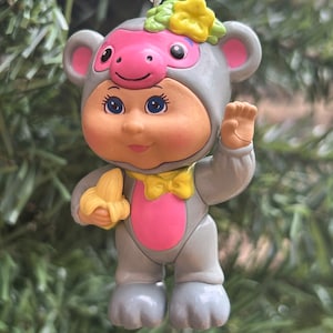 Peut inclure: Ornement fantaisiste représentant une figurine de poupée dans un costume de singe gris, avec un visage et une poitrine roses. La figurine tient une banane jaune et porte un nœud papillon jaune. Une fleur jaune et des feuilles vertes ornent la tête. L'ornement est suspendu à une ficelle blanche.