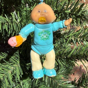 May include: A Cabbage Patch Kids doll ornament with a tan complexion, wearing a light blue long-sleeved shirt with the green Cabbage Patch Kids logo and cream-coloured trousers. The doll has a yellow dummy and holds a pink ice cream cone.