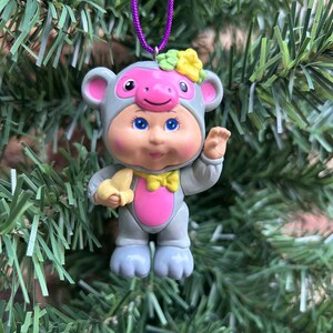 May include: A novelty Christmas tree decoration featuring a baby in a grey monkey outfit. The ornament has a pink face and chest, blue eyes, and a yellow bow tie. It holds a yellow banana and hangs from a purple cord.