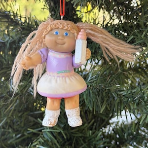 May include: A Cabbage Patch Kids doll ornament with blonde yarn hair in pigtails, wearing a lavender and white dress. The doll has blue eyes, a light skin tone, and holds a small white baby bottle. It hangs from a thin red string.