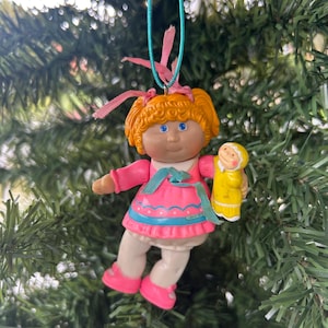 May include: A vintage Cabbage Patch Kids doll Christmas tree ornament. The doll has orange hair, blue eyes, and is wearing a pink dress with a blue trim. The doll is holding a yellow gingerbread man. The ornament is suspended from a green string.