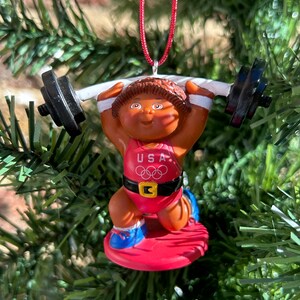 May include: A Christmas tree ornament depicting a weightlifter doll with brown skin, wearing a red USA Olympic uniform and lifting a barbell. The ornament has black weights, a red base, and is suspended by a red string. The background is a green Christmas tree.