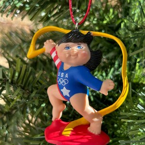 May include: A Christmas tree ornament depicting a gymnast in a blue leotard with a white star and the text "USA" and Olympic rings. The gymnast is on a red platform with a yellow ribbon. The ornament hangs from a red cord.