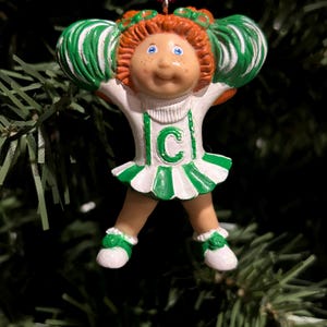 May include: A green and white cheerleader ornament with red hair, blue eyes, and a 'C' on the front. The ornament is hanging from a red string.