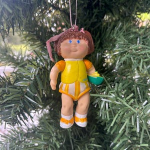 May include: A vintage Cabbage Patch Kids Christmas ornament. The doll is wearing a yellow vest and striped nappy. It has blue eyes and brown hair. The ornament is hanging from a pink ribbon and is holding a small green bowl.