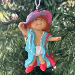 May include: A Christmas tree ornament depicting a doll-like figure with red hair, wearing a purple hat with a pink feather, a blue coat, and red shoes. The doll has a pearl necklace and a red purse.