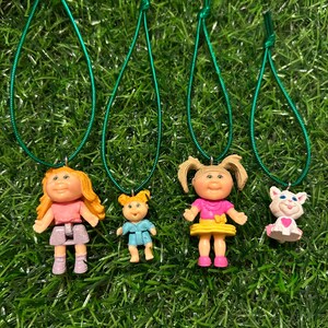 May include: Four Cabbage Patch Kids necklaces with green cords. The necklaces feature a blonde girl with a pink dress, a blonde girl with a blue dress, a blonde girl with a yellow dress, and a white cat with a pink dress.