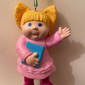 May include: A vintage Cabbage Patch Kids ornament. The doll has blonde pigtails, blue eyes, and a pink dress. It holds a blue book and wears pink boots. A green string is attached for hanging, suitable for a Christmas tree.
