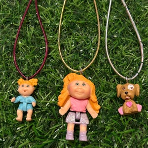 Puede incluir: Tres collares con diferentes dijes. El primer collar tiene un cordón rojo y un pequeño dije de muñeca Cabbage Patch Kid vestida de azul. El segundo collar tiene una cadena dorada y un dije de muñeca Cabbage Patch Kid con una camisa rosa y una falda plateada. El tercer collar tiene una cadena plateada y un dije de perro marrón con un corazón rosa.