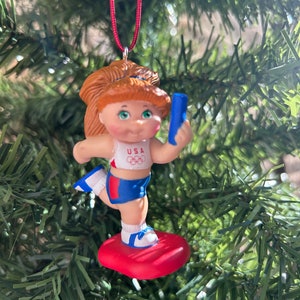 May include: A Christmas tree ornament depicting a doll-like figure in a USA Olympic uniform, holding a blue baton. The figure has red hair and stands on a red base. The ornament is suspended from a red cord.