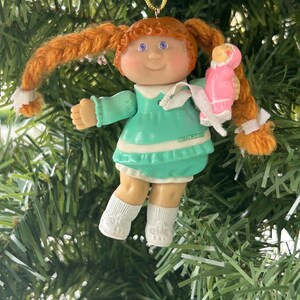 May include: A vintage Cabbage Patch Kids Christmas ornament. The doll has orange yarn pigtails, blue eyes, and a green dress. It holds a small pink doll. The ornament is suspended from a gold string.