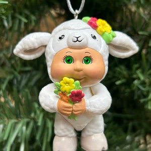 May include: A white lamb-themed ornament with a smiling face, green eyes, and a floral headband. The ornament holds a bouquet of yellow and red flowers. It is hanging from a silver string, perfect for holiday decor.