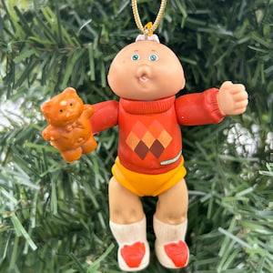 May include: A Cabbage Patch Kids Christmas tree ornament. The doll has a peach-coloured face, blue eyes, and a red jumper with an argyle pattern. It holds a small brown teddy bear. The doll is wearing yellow shorts and white socks with red hearts.