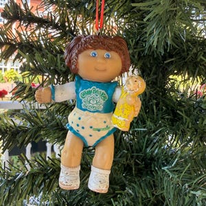 May include: A Cabbage Patch Kids doll ornament with brown hair, blue eyes, and a teal top featuring the brand logo. The doll wears white polka-dot bottoms and white shoes, and holds a small yellow toy. A red ribbon is attached for hanging.