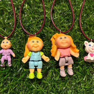May include: Four pendant necklaces featuring small plastic dolls and a cat. The dolls have blonde or light-coloured hair and wear colourful outfits. The cat pendant is white with a pink heart. The necklaces have maroon cords, set against a green background.