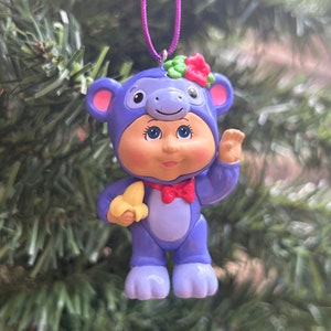 Peut inclure: Ornement de Noël en forme de singe de dessin animé violet et blanc, portant une capuche et un nœud papillon, tenant une banane. L'ornement a une fleur rouge sur la tête et est suspendu à une ficelle violette. L'arrière-plan est un sapin de Noël flou.