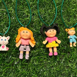 May include: Four pendant necklaces with green cords. The pendants feature a white cat with a pink heart, a doll with orange hair, a doll with black pigtails, and a doll with blonde pigtails. The dolls wear pink, yellow, and blue outfits.
