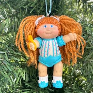 May include: A Cabbage Patch Kids doll Christmas tree ornament. The doll has orange yarn pigtails, blue eyes, and a blue and white striped outfit. It holds a yellow comb and hangs from a blue ribbon against green Christmas tree branches.