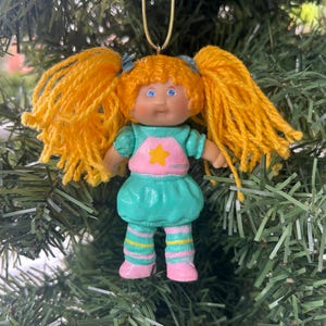 May include: A vintage doll Christmas tree ornament. The doll has yellow yarn pigtails, blue eyes, and a pink and teal outfit. It features a star on the dress and hangs from a gold string, ideal for festive decor.