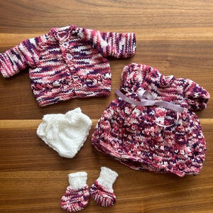 May include: A knitted baby doll outfit in shades of pink, purple, and white. The set includes a cardigan, dress, nappy cover, and booties.