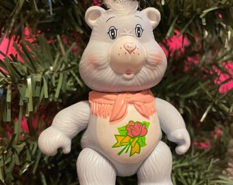 May include: A light blue Care Bear ornament with a pink scarf and a rose design on its belly. The bear has a white tuft of hair and is hanging from a silver string. The ornament is set against a green Christmas tree.