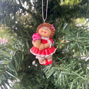 May include: A Cabbage Patch Kids Christmas tree decoration. The ornament depicts a doll with brown curly hair, blue eyes, and a pink bow. The doll wears a red dress and holds a pink ice cream cone. It hangs from a white string.