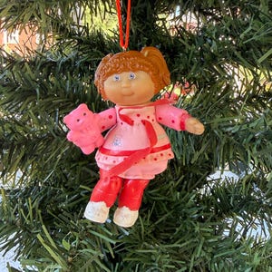 May include: A hanging doll ornament with auburn hair in pigtails, wearing a pink dress with red polka dots and a small bear graphic. The doll has red trousers, white shoes, and holds a small pink bear figure in one hand, suspended by a red ribbon.