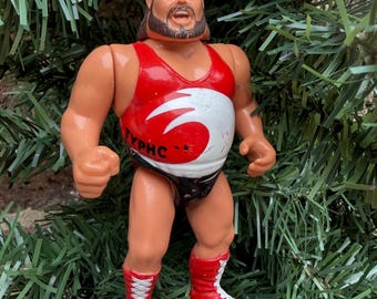 May include: A plastic action figure of a wrestler, with a red and white top, black trunks, and red boots. The figure has a brown beard and hair. The top has the word "TYPHO" printed on it. The figure is hanging from a green string.