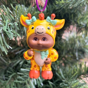 May include: A charming ornament depicting a baby doll dressed as a giraffe. The doll has brown skin and is wearing a yellow giraffe hood with orange spots and green bows. The ornament hangs from a purple cord and is set against a backdrop of green Christmas tree branches.