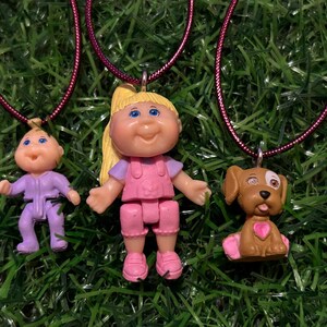 May include: Three pendant necklaces featuring Cabbage Patch Kids and a dog. The pendants are attached to a dark pink cord. The Cabbage Patch Kids are wearing pink and purple outfits. The dog pendant has a pink heart.