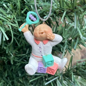 May include: A baby-themed Christmas ornament. The ornament depicts a seated baby in a white onesie with a pink collar and purple buttons. The baby holds a rattle and sits on three colourful blocks. The ornament hangs from a silver chain.